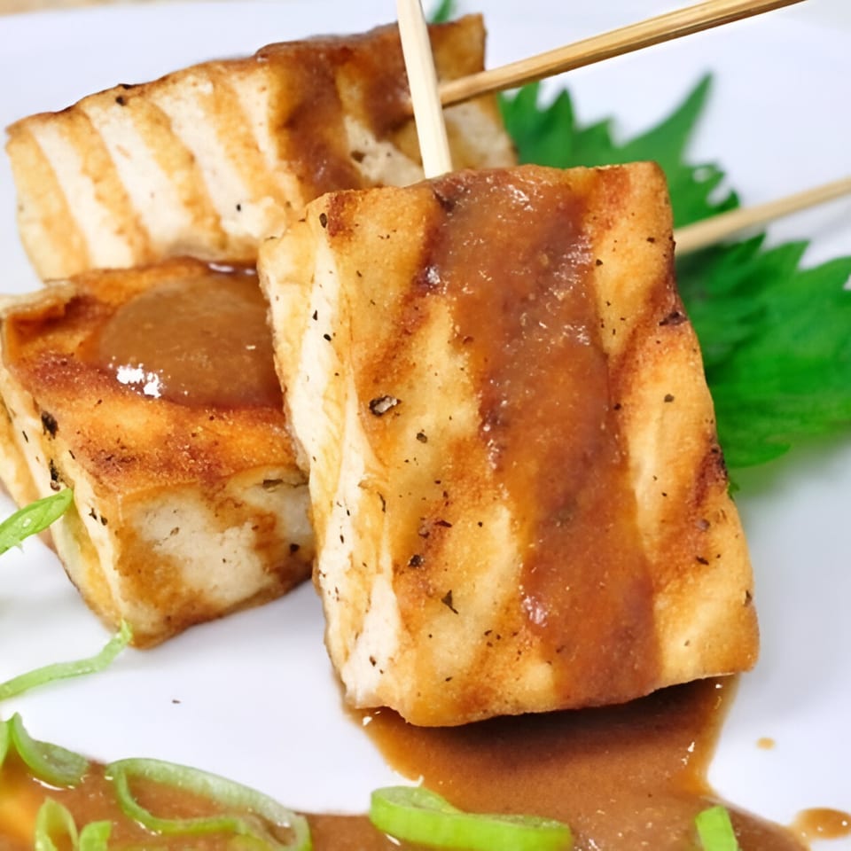 Grilled tofu skewers with a rich brown sauce and green garnish on a white plate.